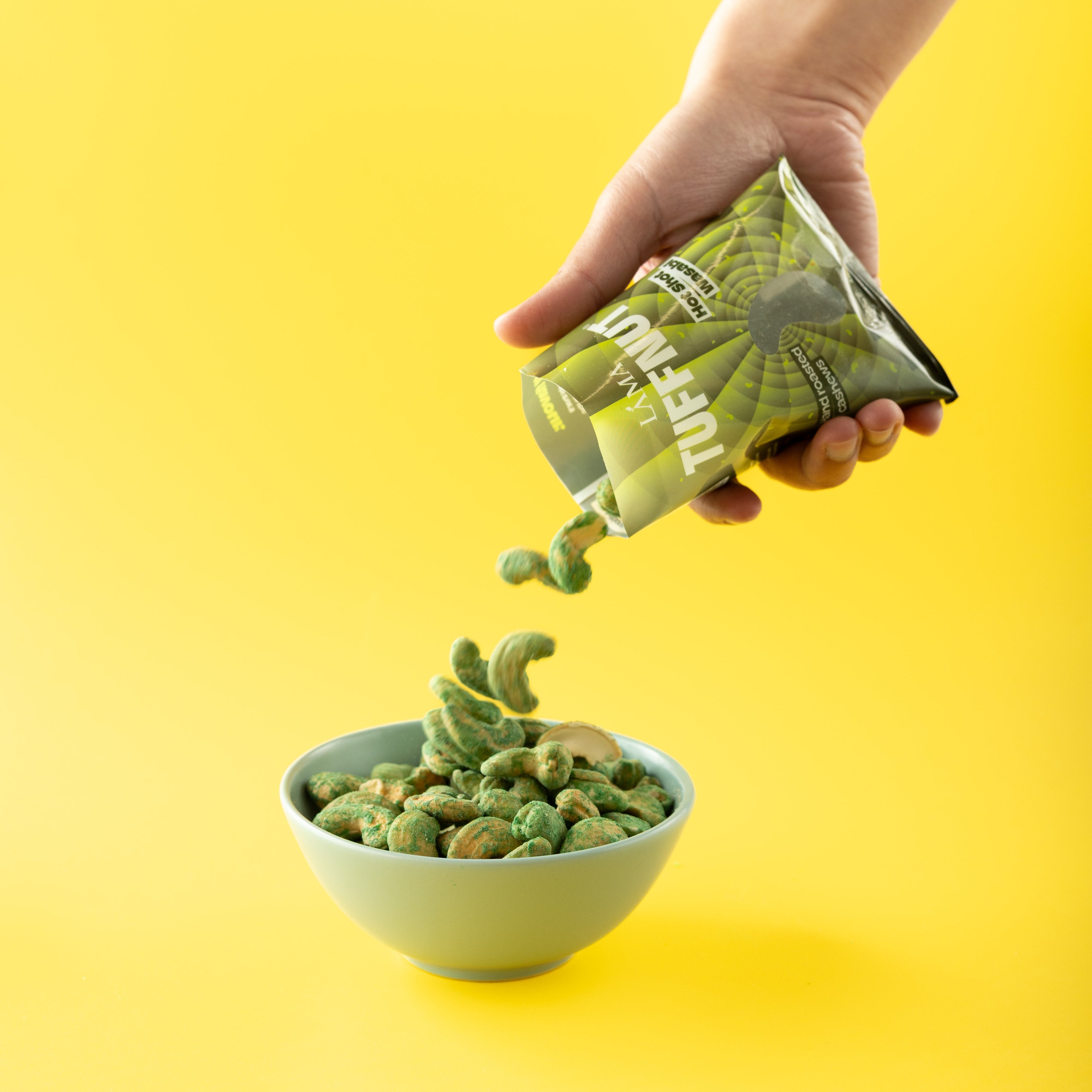 Hand pouring TuffNut Wasabi Cashews from a bag into a bowl against a yellow background