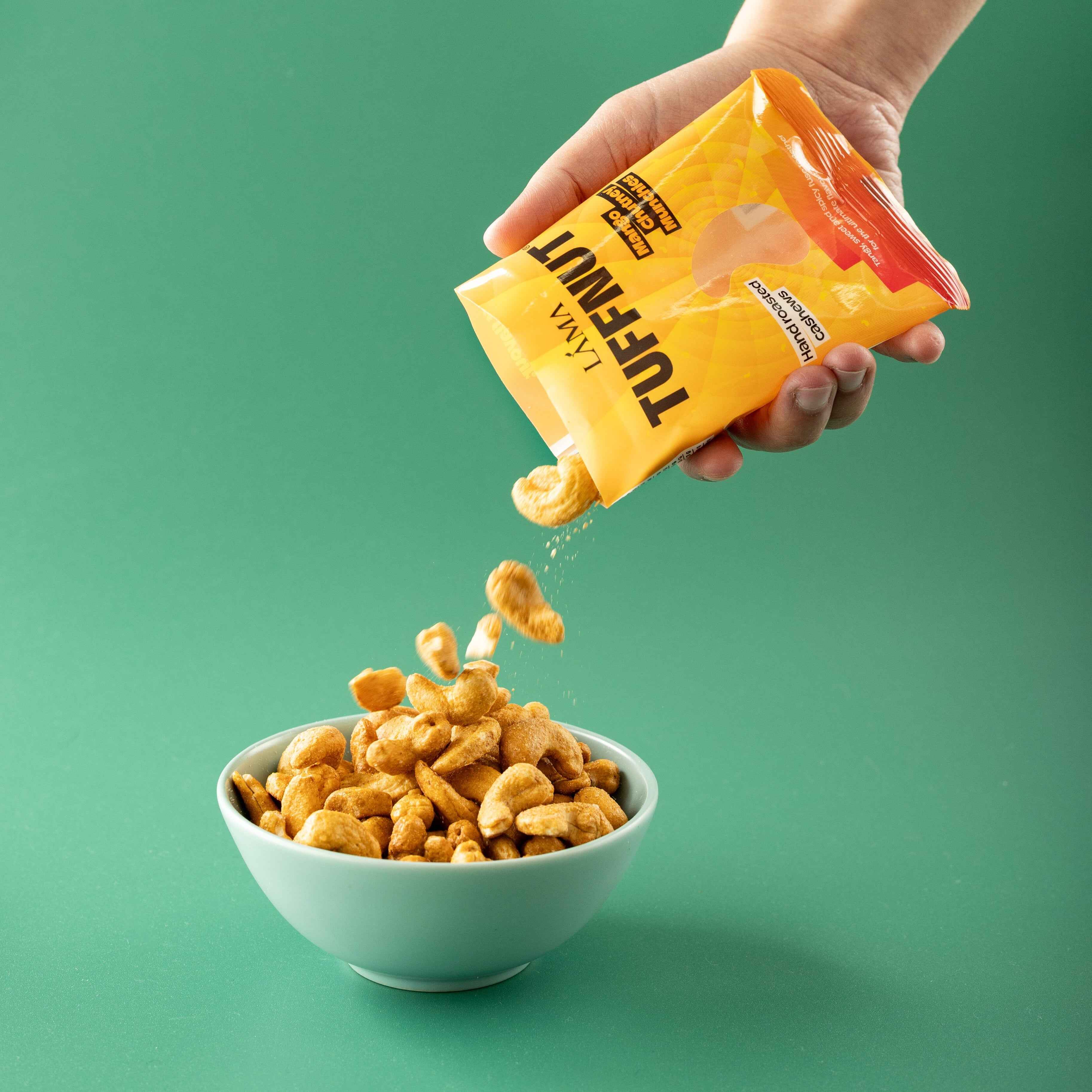 Hand pouring TuffNut Mango Chutney Munchies Hand Roasted Cashews into a bowl against a green background