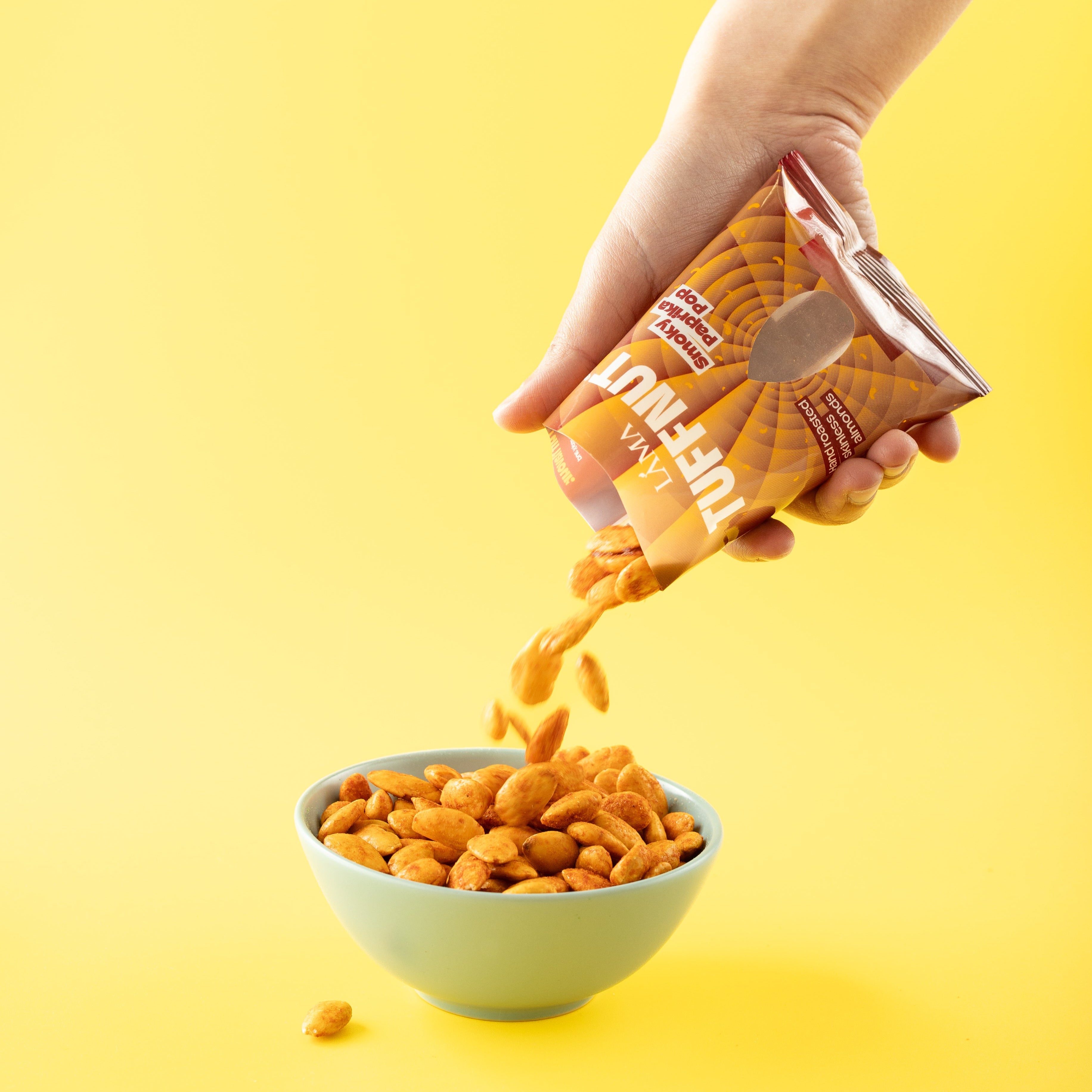 Hand pouring smoky paprika pop tuffnut almonds snack from a bag into a bowl on a yellow background